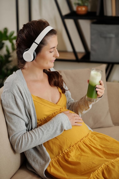 Kobieta w ciąży słuchająca muzyki w słuchawkach i pijąca zielony koktajl