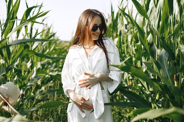 Kobieta w ciąży spaceru w polu kukurydzy. Brunetka kobieta spacerująca w letnim polu nosząca białe ubrania