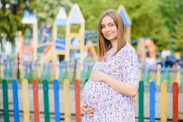 Kobieta W Ciąży Stoi W Parku Obok Placu Zabaw, Na Którym Bawią Się Dzieci.