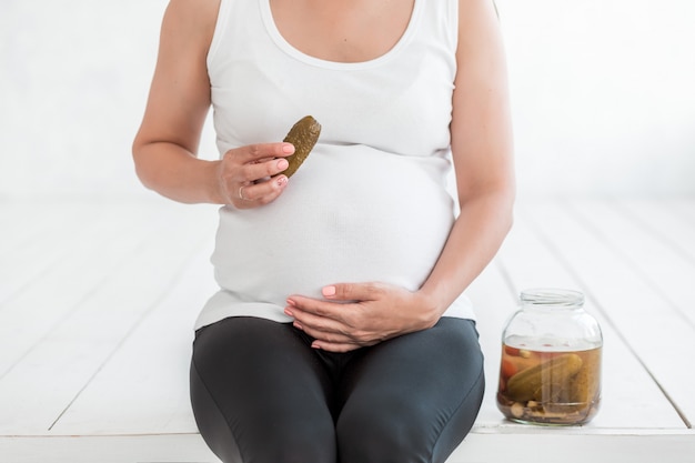 Kobieta w ciąży trzyma solone ogórki w słoiku w pobliżu jej brzucha.