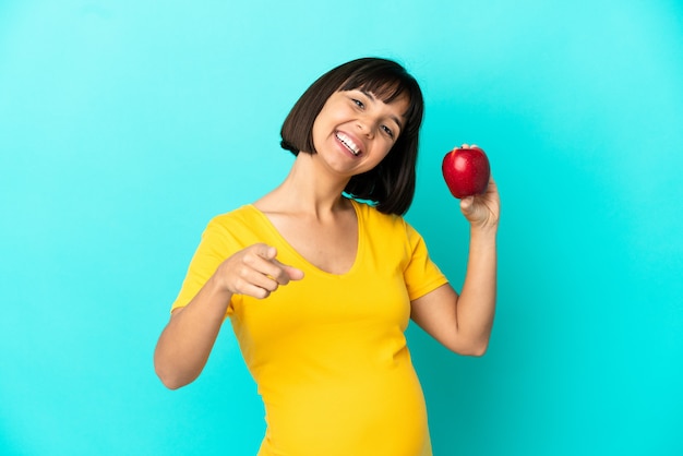 Kobieta w ciąży trzymająca jabłko na białym tle na niebieskim tle wskazująca przód ze szczęśliwym wyrazem twarzy