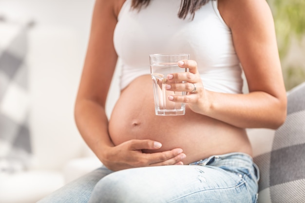 Kobieta w ciąży trzymająca się jedną ręką za brzuch, w drugiej trzyma szklankę wody.