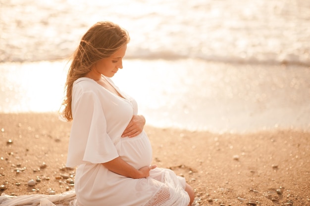 Kobieta w ciąży ubrana w białą sukienkę siedząca nad morzem