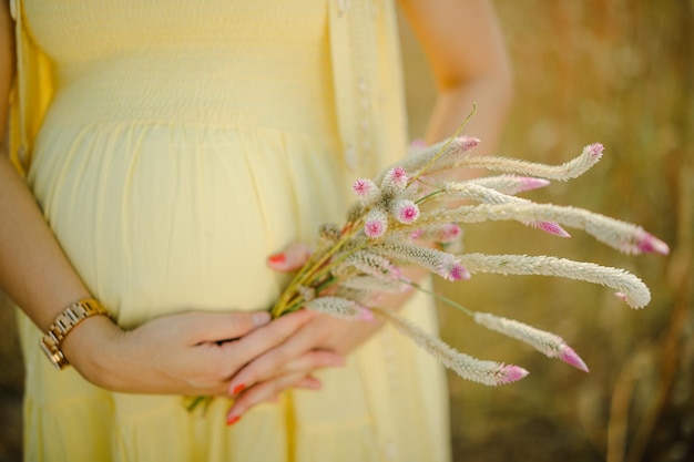 Kobieta w ciąży, ubrana w jasnożółtą sukienkę, trzymająca w rękach bukiet kwiatów stokrotek.