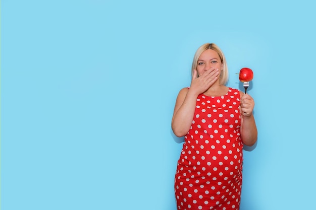 Kobieta w ciąży w czerwonej sukience w kropki z jabłkiem na widelcu w dłoni zamyka usta drugą ręką na niebieskim tle z copyspace koncepcja żywienia dla kobiet w ciąży