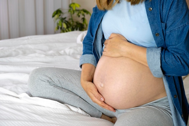 Kobieta w ciąży w niebieskiej koszulce i dżinsowej koszuli siedzi w łóżku z rękami na brzuchu Ciąża koncepcja przygotowania i oczekiwania na macierzyństwo Miłość opieka nad dzieckiem i rodzicielstwo