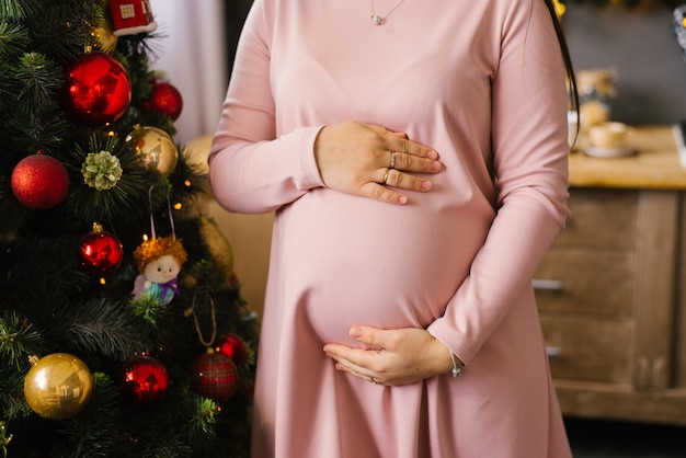 Kobieta w ciąży w różowej sukience stoi obok choinki i trzyma ręce na brzuchu