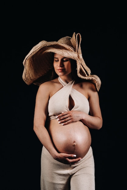 Kobieta w ciąży w słomkowym kapeluszu w beżowych ubraniach w studio na czarnym tle.