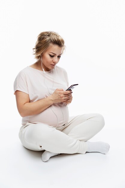 Kobieta w ciąży w słuchawkach ze smartfonem w dłoniach.