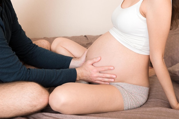 Kobieta w ciąży z mężem siedzącym na łóżku trzymając ręce za brzuch w domu