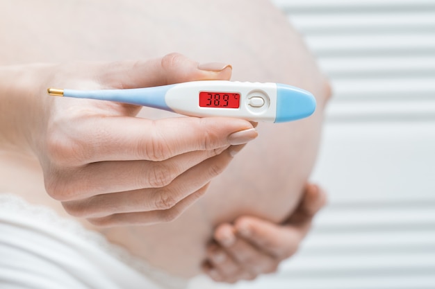 Kobieta w ciąży z termometrem w ręku i wysoką temperaturą. Choroby podczas koncepcji ciąży.