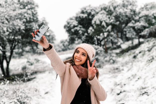 Kobieta W Ciepłej Odzieży Wierzchniej Stojącej W Lesie I Robienia Selfie Malowniczego śnieżnego Krajobrazu Podczas Korzystania Ze Smartfona