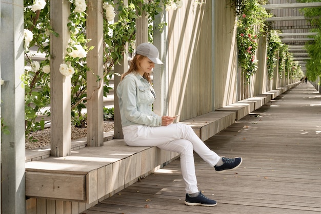 Kobieta w codziennych ubraniach siedzi na drewnianej ławce z białymi różami na ścianie na zewnątrz