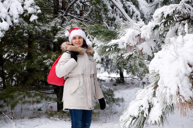 Kobieta w czapce Mikołaja spaceruje po zaśnieżonym lesie sosnowym