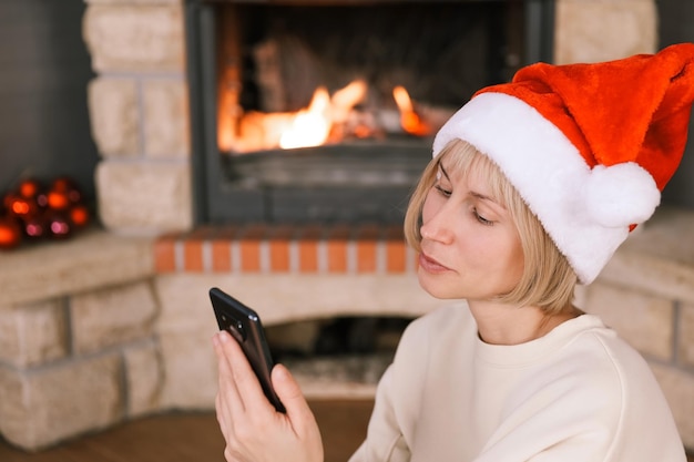 Kobieta w czapce Świętego Mikołaja komunikuje się przez telefon przy płonącym kominku w przytulnym domu