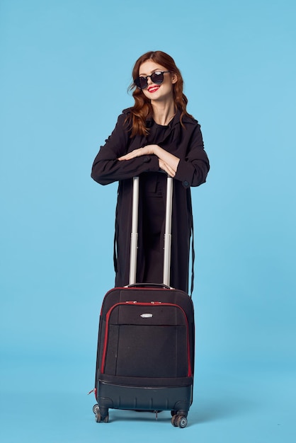 Kobieta w czarnym płaszczu walizka okulary wakacje niebieskie tło