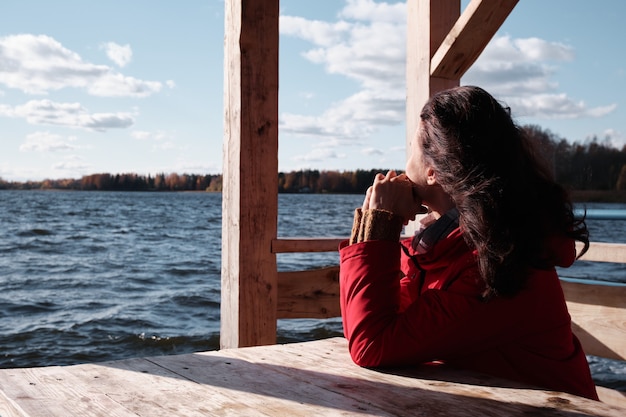 Kobieta w czerwonej kurtce siedzi przy stoliku na drewnianym molo i patrzy na wodę