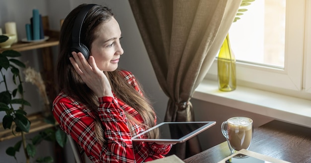 Kobieta w czerwonej sukience słucha audiobooka w słuchawkach z tabletem przy oknie Koncepcja nowoczesnej edukacji technologicznej i studiowania książek z radością