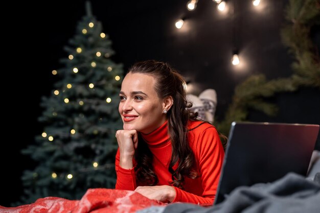Kobieta w czerwonym swetrze leżąc na łóżku w pobliżu swojego laptopa i odwracając wzrok, pozując do kamery. Szczęśliwego Nowego Roku i wesołych świąt zdjęcie koncepcji