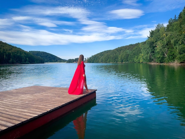 Kobieta w długiej czerwonej sukience stojąca na drewnianym pontonie w pobliżu jeziora otoczonego lasem