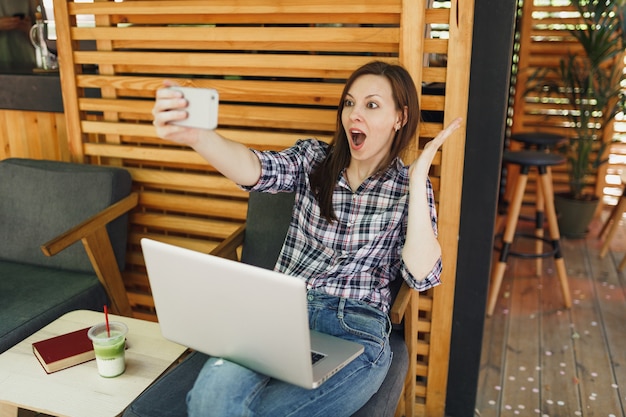 Kobieta W Drewnianej Kawiarni Na Zewnątrz Ulicy Lato Siedzi Z Laptopa Komputer Pc, Robi Selfie Strzał Na Telefon Komórkowy, Relaks W Czasie Wolnym. Biuro Mobilne