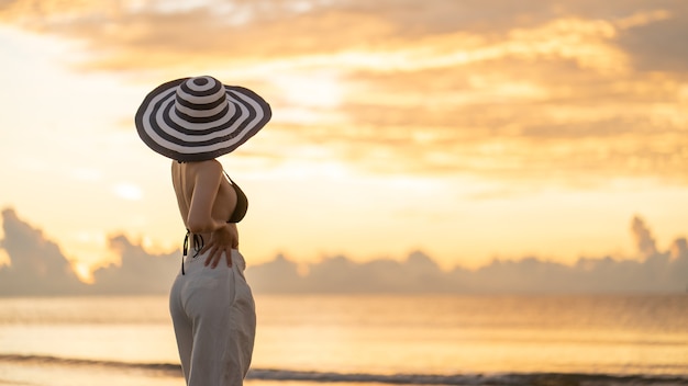 Kobieta w górnym bikini i białe długie spodnie w kapeluszu na plaży z pięknym wschodem słońca