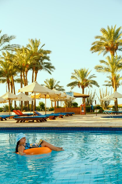 Kobieta w kapeluszu przeciwsłonecznym w basenie w kręgu pływackim z laptopem na tle palm i leżaków.