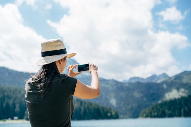 Kobieta w kapeluszu robi zdjęcia gór swoim telefonem z tyłu