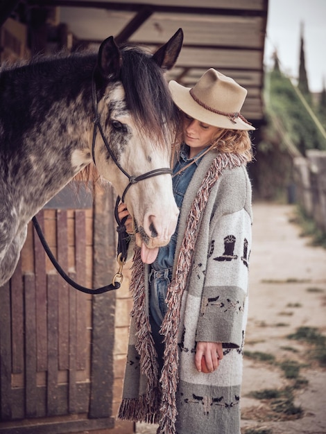 Kobieta w kapeluszu stojąca w pobliżu uroczego konia z cętkowaną sierścią i uzdą stojąca w stajni w letni dzień na wsi