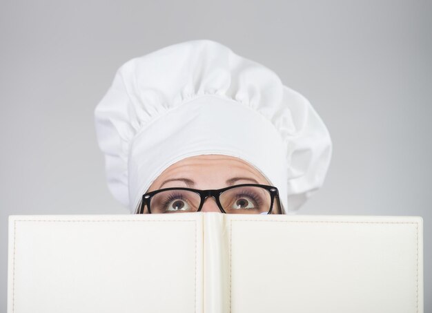 Kobieta w kapeluszu szefa kuchni patrząca w kamerę nad książką kucharską