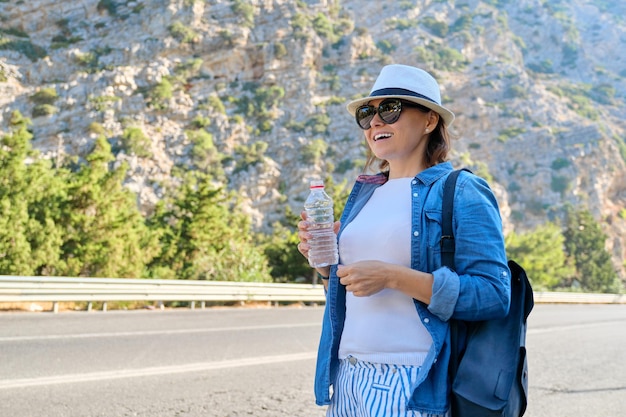 Kobieta w kapeluszu z butelką wody w górach na autostradzie, autostop, zatrzymanie autobusu, z kopią miejsca. Natura, droga, turystyka, podróże, przygoda, tło koncepcji wycieczki samochodowej