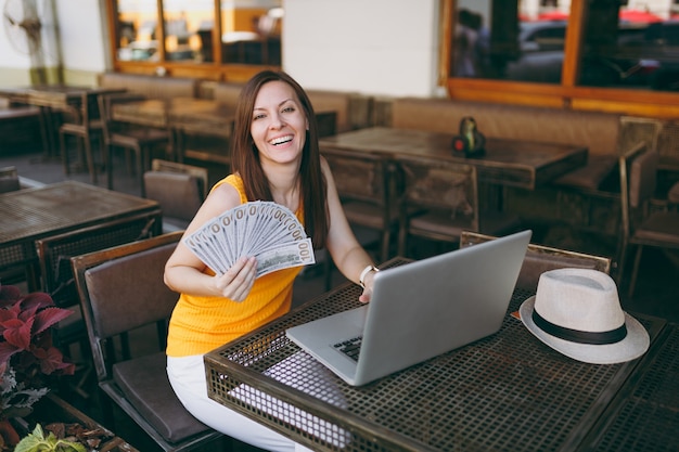 Kobieta W Kawiarni Na świeżym Powietrzu W Kawiarni Siedząc Z Nowoczesnym Komputerem Typu Laptop, Trzyma W Ręku Kilka Banknotów Dolarów