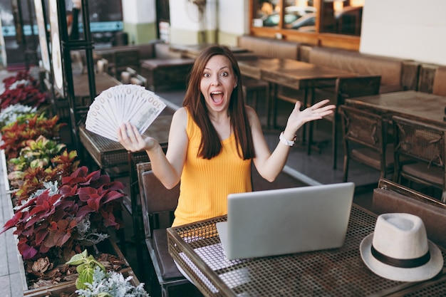 Kobieta W Kawiarni Na świeżym Powietrzu W Kawiarni Siedząc Z Nowoczesnym Komputerem Typu Laptop, Trzyma W Ręku Kilka Banknotów Dolarów