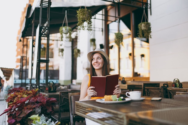 Kobieta W Kawiarni Na Zewnątrz Ulicy Kawiarnia Siedzi Przy Stole W Kapeluszu, Czyta Książkę Z Filiżanką Cappuccino, Ciasto, Relaks W Restauracji W Czasie Wolnym