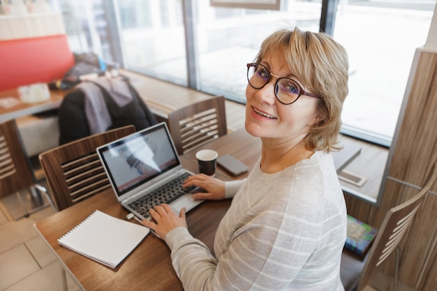 Kobieta w kawiarni pracuje na laptopie. Ona się uśmiecha. Kobieta w średnim wieku, dorosła osoba w biurze. Praca zdalna.