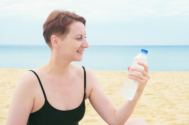 Kobieta w kostiumie kąpielowym siedzi na plaży i trzyma w dłoni plastikową butelkę wody pitnej