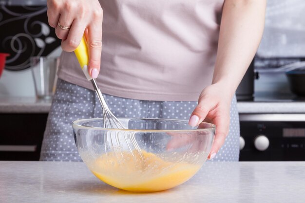 Kobieta w kuchni trzyma w dłoniach trzepaczkę z żółtą rączką i ubija je jajkami w szklanej przezroczystej misce Codzienna rutyna matki, która robi jajecznicę na śniadanie dla rodziny