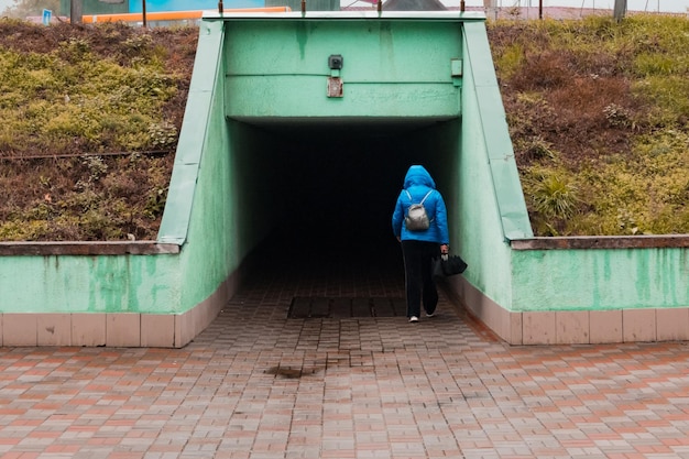 Kobieta w kurtce podczas deszczu wchodzi do tunelu pod drogą z parasolem w miejskim mieście Kałuża Architektura Miasto Korytarz Ochrona Pogoda poniżej Kropla deszczu Widok z tyłu Miejski