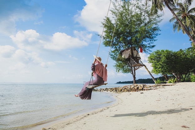 Kobieta w letnim kapeluszu na plaży, ciesząc się widokiem na ocean, jeżdżąc na huśtawce w Karimun Jawa