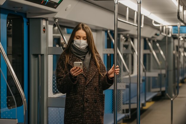 Kobieta w medycznej masce na twarz, aby uniknąć rozprzestrzeniania się koronawirusa, stoi i korzysta ze smartfona w pustym wagonie metra. Dziewczyna w masce chirurgicznej przewija wiadomości na swoim telefonie komórkowym w pociągu.