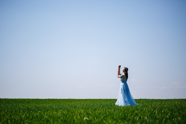 Kobieta w niebieskiej długiej sukni na tle zielonego pola. Moda piękny portret pięknej dziewczyny z uśmiechem na twarzy