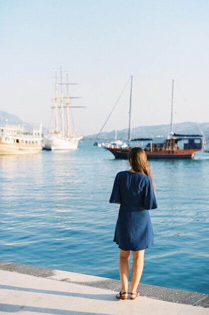 Kobieta w niebieskiej sukience stoi na molo i patrzy na statki