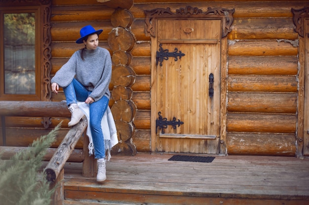 Kobieta w niebieskim kapeluszu i swetrze siedzą jesienią przy drzwiach drewnianego domu z bali