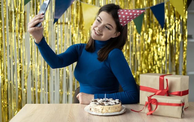Kobieta w odświętnej czapce z uśmiechem na twarzy siedzi na urodziny na imprezie online za pomocą smartfona i aplikacji Rozmowa wideo ze znajomymi na odległość
