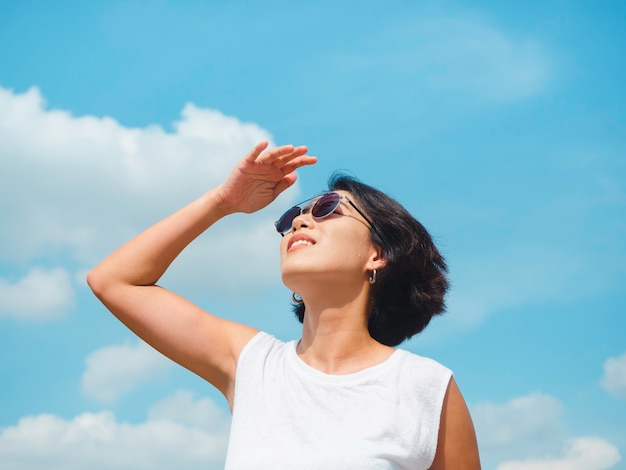 Kobieta w okresie letnim. Uśmiechnięta piękna Azjatycka kobieta z krótkimi włosami w okularach przeciwsłonecznych i białej koszuli bez rękawów, patrząc w górę i ocieniając oczy ręką na tle błękitnego nieba w słoneczny dzień lata.