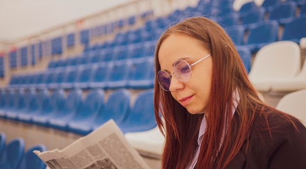 Kobieta w okularach i garniturze czyta gazetę siedząc na stadionie