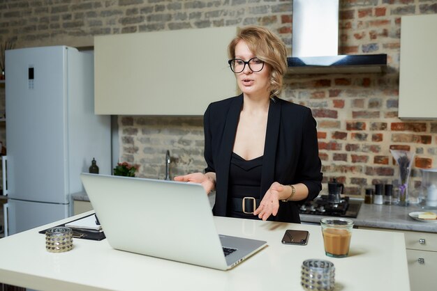 Kobieta W Okularach Pracuje Zdalnie Na Laptopie W Kuchni. Blond Dziewczyna Gestykuluje, Rozmawia Z Kolegami Na Odprawie Biznesowej W Domu.