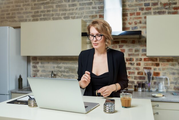 Kobieta w okularach pracuje zdalnie na laptopie w kuchni. Dziewczyna rozmawia ze swoimi kolegami na odprawie biznesowej online w domu.