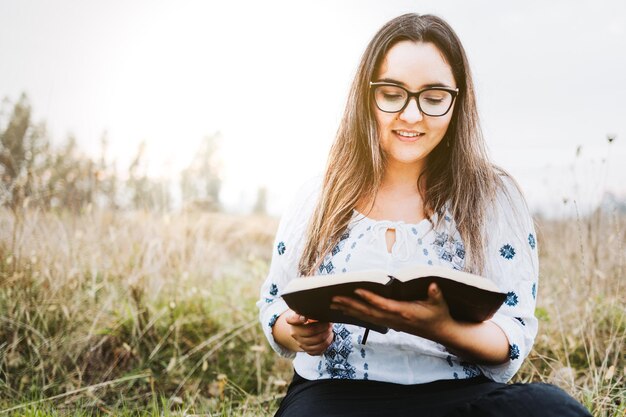 Kobieta w okularach siedzi na trawie na świeżym powietrzu, trzymając i czytając otwartą Biblię Światło słoneczne