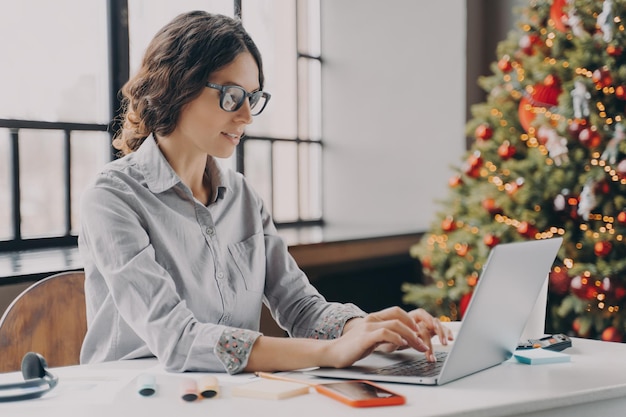 Kobieta W Okularach Siedzi W Biurze W Pobliżu Choinki I Pracuje Na Laptopie